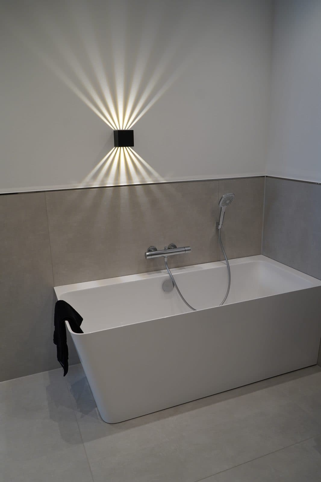 Freistehende weiße Rechteckwanne vor beigem Fliesenspiegel unter einer Wandleuchte mit Lichtfächer, Partner-Ausstellung ELEMENTS Neustadt
