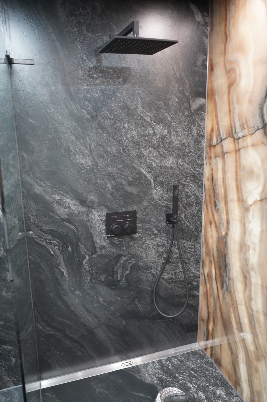 Dusche mit Regen- und Handbrause, dunkler Naturstein neben einer Onyxwand