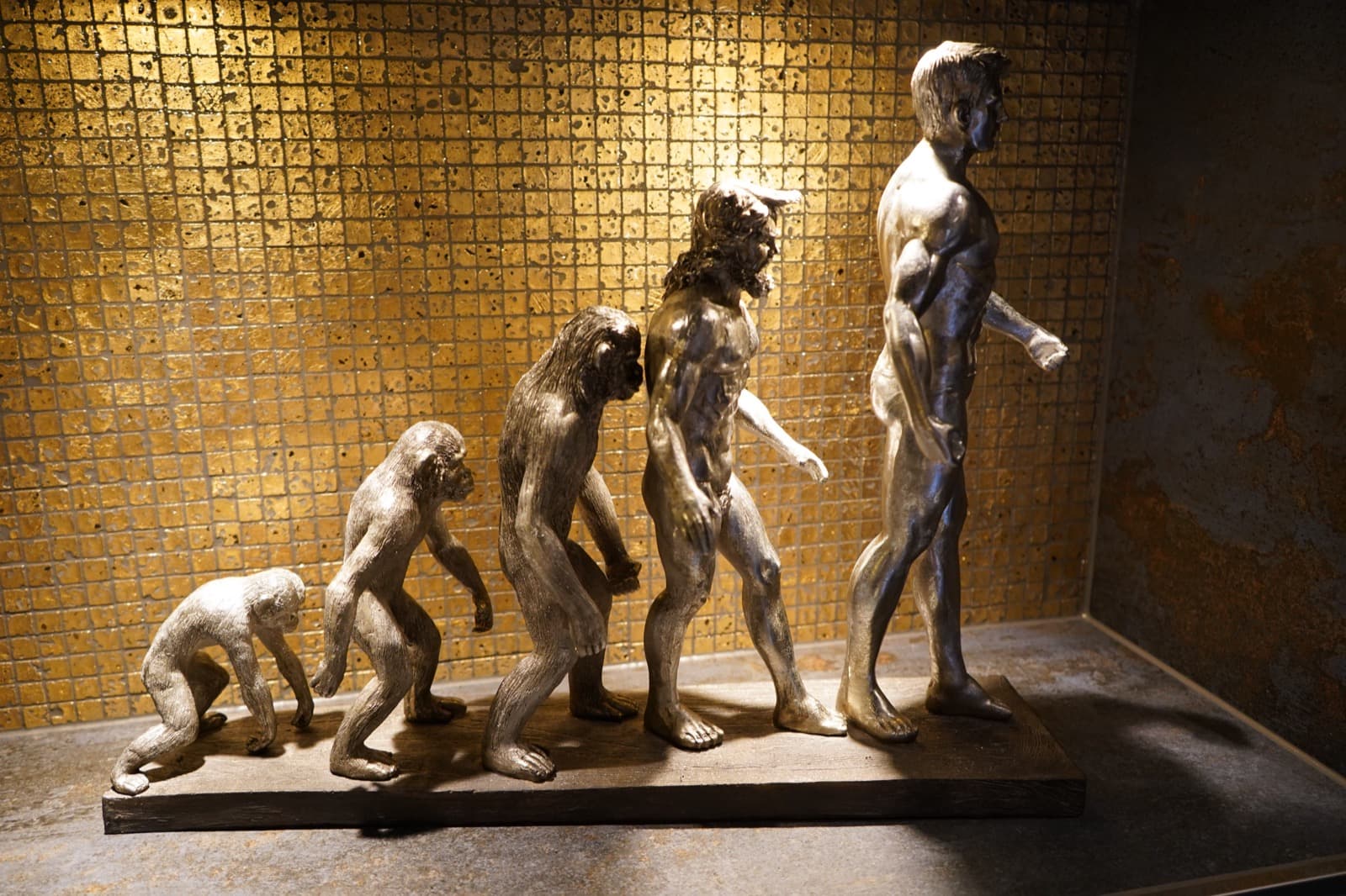 Skulpturenreihe vom Affen zum Menschen vor einer goldenen Mosaikwand